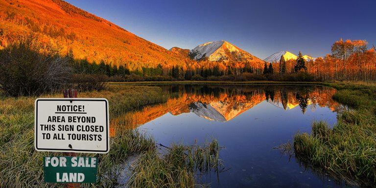 Mountain landscape with autumn foliage reflected in a calm lake. A sign in the foreground reads "Area beyond this sign closed to all tourists. For sale land.