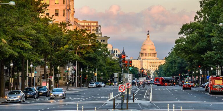 A view of a wide city street lined with trees and buildings, with the U.S. Capitol building visible in the background under a partly cloudy sky.
