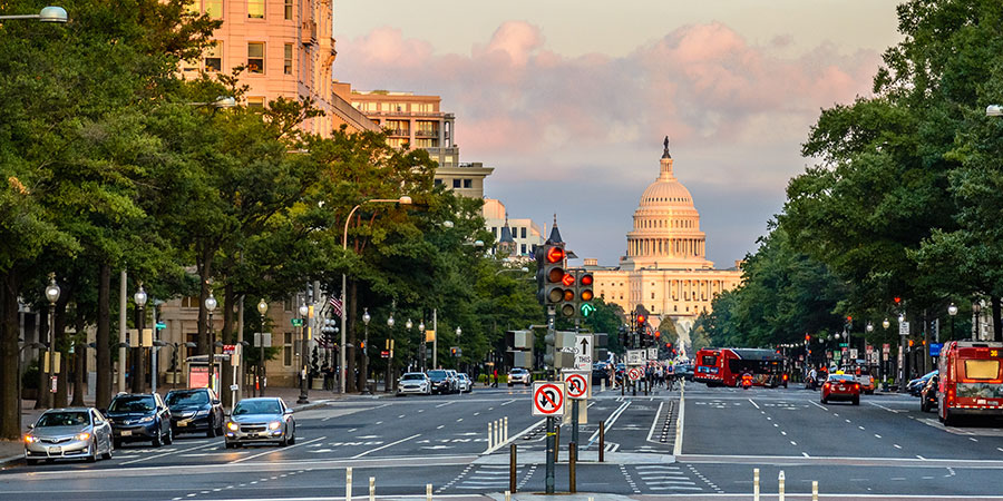 A view of a wide city street lined with trees and buildings, with the U.S. Capitol building visible in the background under a partly cloudy sky.