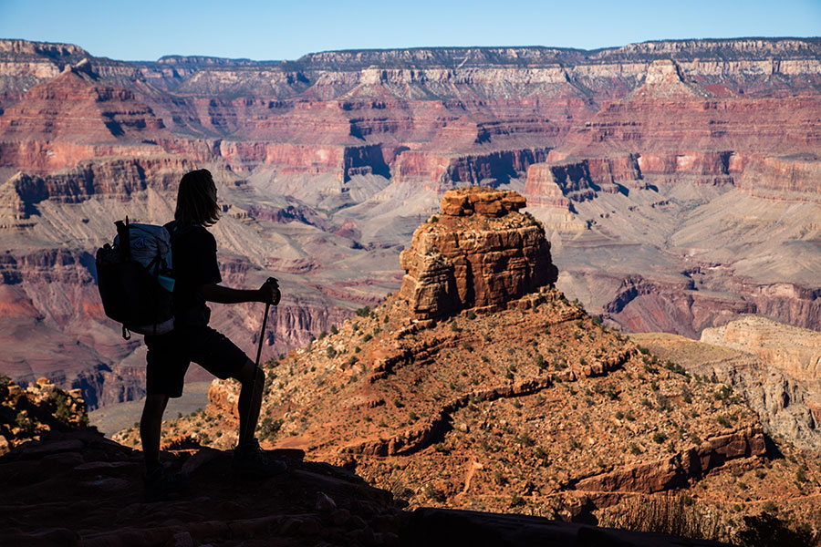 A hiker with a backpack stands on a rocky ledge, looking out over the layered rock formations of the Grand Canyon under a clear sky.