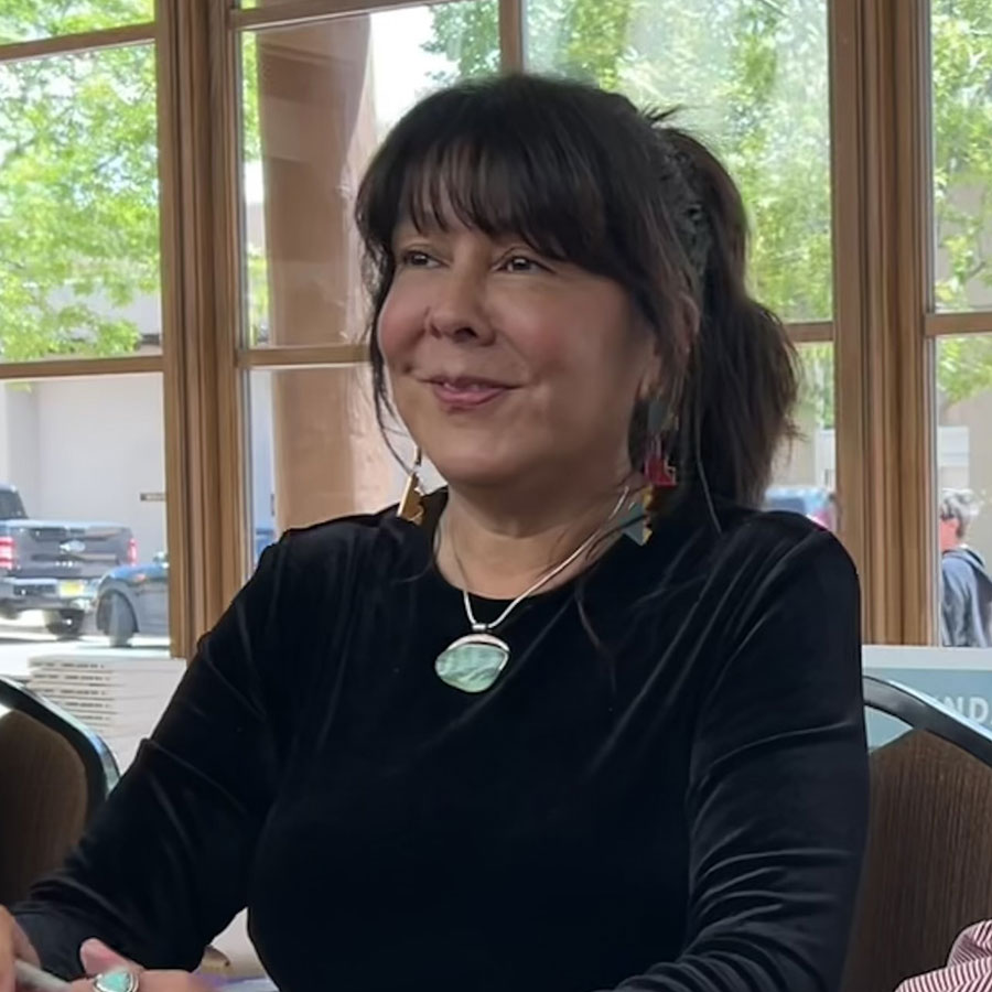 Deborah Jackson Taffa, a woman with dark hair in a ponytail, wearing a black top and a necklace, sits indoors in front of large windows with trees visible outside.