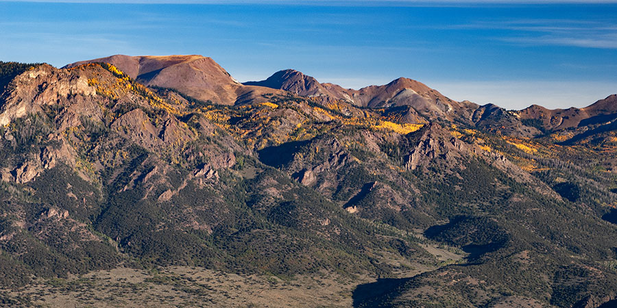Mountain range with rugged peaks and patches of yellow and green vegetation under a clear blue sky.