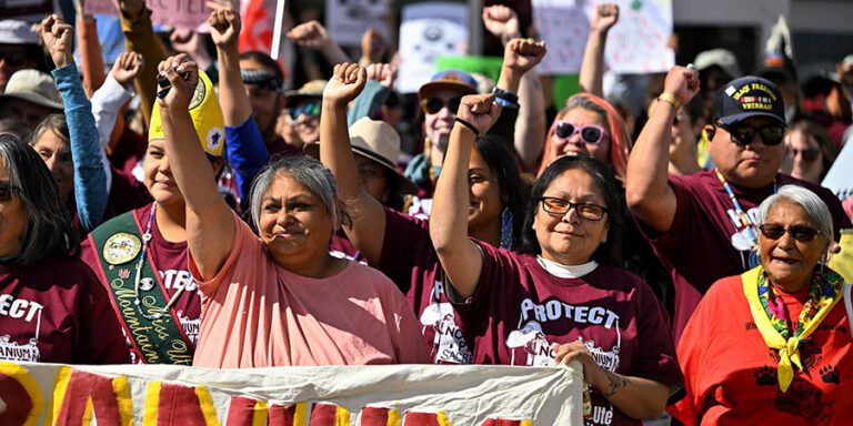 A group of tribal members at a protest rally hold their fists up and stand behind a banner, some wearing matching maroon shirts that read "PROTECT White Mesa," as they voice opposition to a uranium mill.