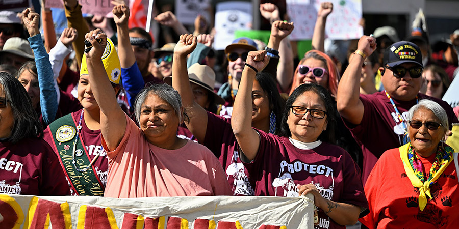 A group of tribal members at a protest rally hold their fists up and stand behind a banner, some wearing matching maroon shirts that read "PROTECT White Mesa," as they voice opposition to a uranium mill.