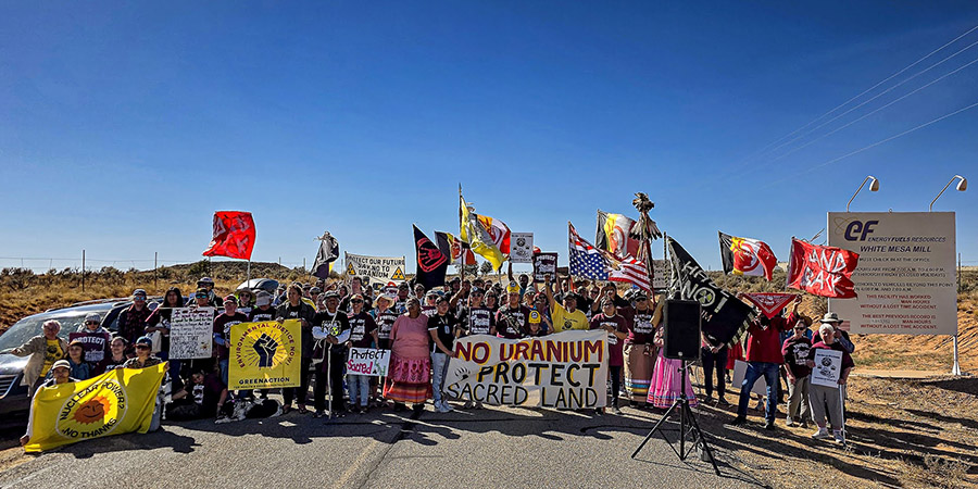 A large group of protesters stand on a road holding signs and flags opposing a uranium mill and mining, with a sign reading “No Uranium Protect Sacred Land” prominent in front.