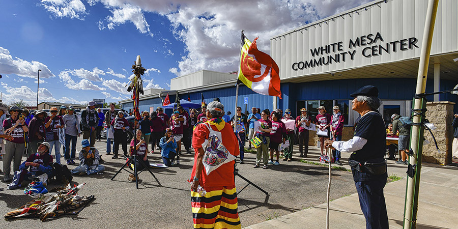 A group of people gathers outside the White Mesa Community Center, some holding flags and wearing traditional clothing, as a person speaks into a microphone about concerns regarding the nearby uranium mill.