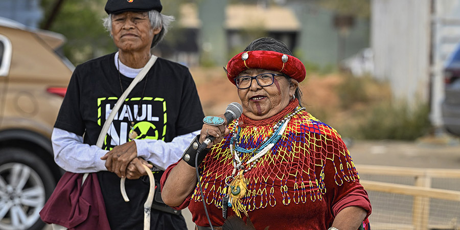 Two Indigenous elders stand outdoors; Dianna Sue White Dove Uqualla, a Havasupai woman, speaks into a microphone about the uranium industry, wearing traditional jewelry and a red hat, while the other stands beside her holding a cane.