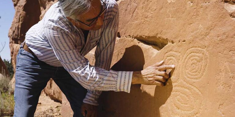 Jim Enote examines petroglyphs on a large rock wall outdoors.