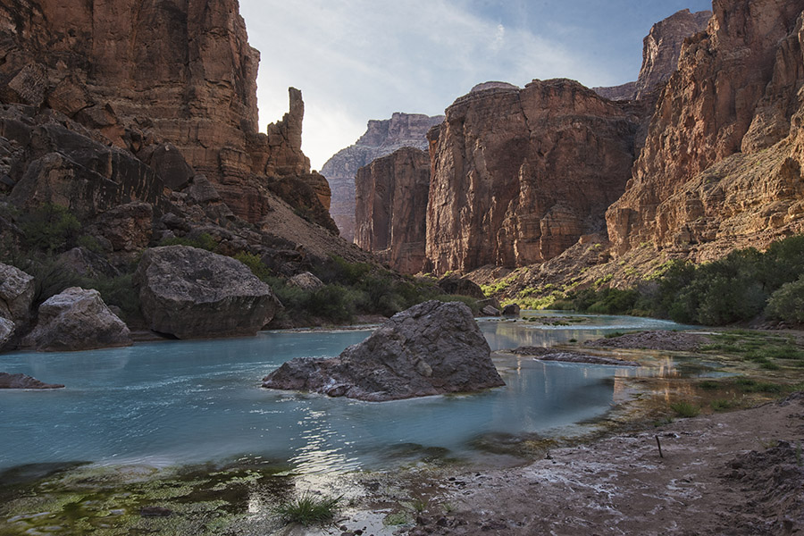 A turquoise river, the Little Colorado River, flows through a rocky canyon with steep red cliffs under a partly cloudy sky.