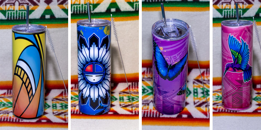Four travel tumblers with Native-inspired and nature-themed designs, featuring abstract patterns, a feathered motif, a butterfly, and a hummingbird, displayed on a patterned blanket.