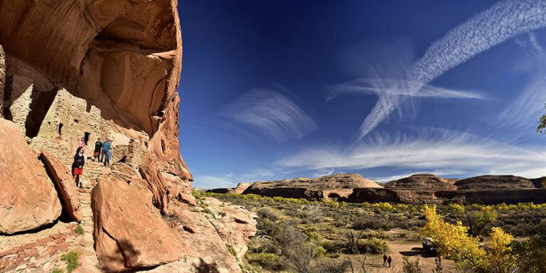 Several people visit cliff dwellings built into a red rock wall, with a vast desert landscape and streaked clouds in the sky.