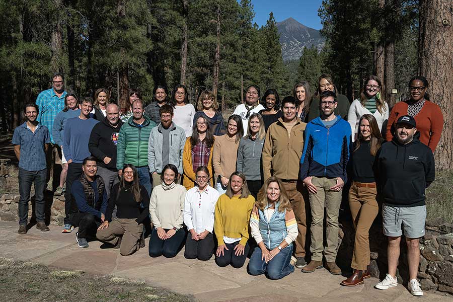 Our Team of 33 people poses for a photo outdoors on a stone path, surrounded by tall pine trees and majestic mountains in the background.