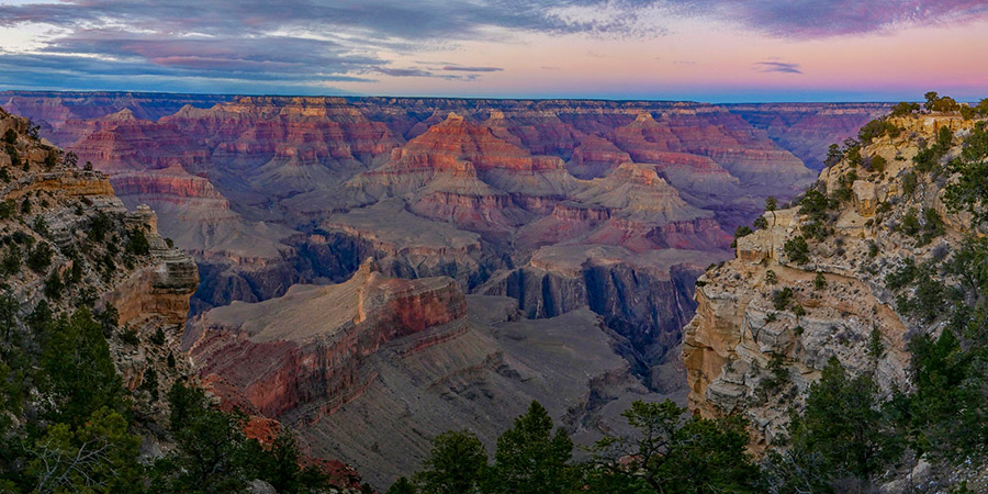 View of the Grand Canyon at sunset, showcasing layered rock formations, steep cliffs, scattered vegetation beneath a partly cloudy sky.