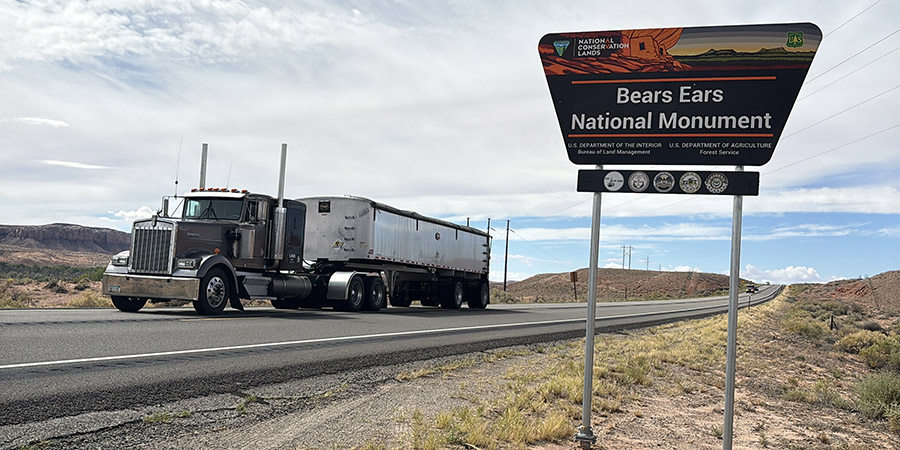 A semi-truck drives on a highway past a roadside sign for Bears Ears National Monument in a desert landscape.