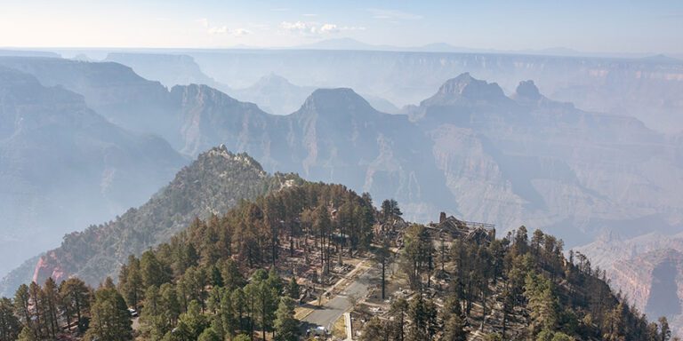 Aerial view of a forested area on a plateau with smoke or mist partially obscuring the layered grand canyon landscape in the background.