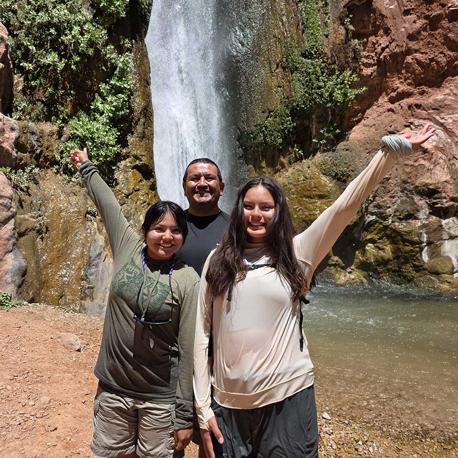 Three people stand in front of a waterfall on a rocky terrain, smiling with arms raised, wearing outdoor clothing.