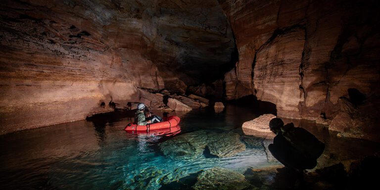 Two people explore a partially flooded cave; one sits in a red inflatable raft on clear blue Grand Canyon water while the other crouches on a rocky ledge.