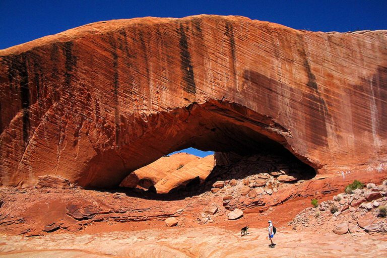 A person and a dog stand beneath a large natural red rock arch with streaked walls, under a clear blue sky in a desert landscape.