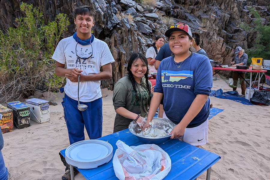 Three people prepare food at an outdoor table on sandy ground, with one person mixing in a large bowl. Other people and supplies are visible in the background near rocks and vegetation.