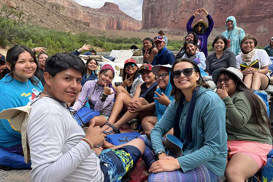 A group of people sit together outdoors on a sunny day in a canyon, smiling and posing for the camera, surrounded by rocky cliffs and greenery.
