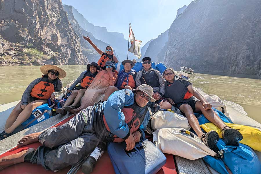 A group of people wearing life jackets and hats sit on a raft with gear as they travel down a river surrounded by steep canyon walls.