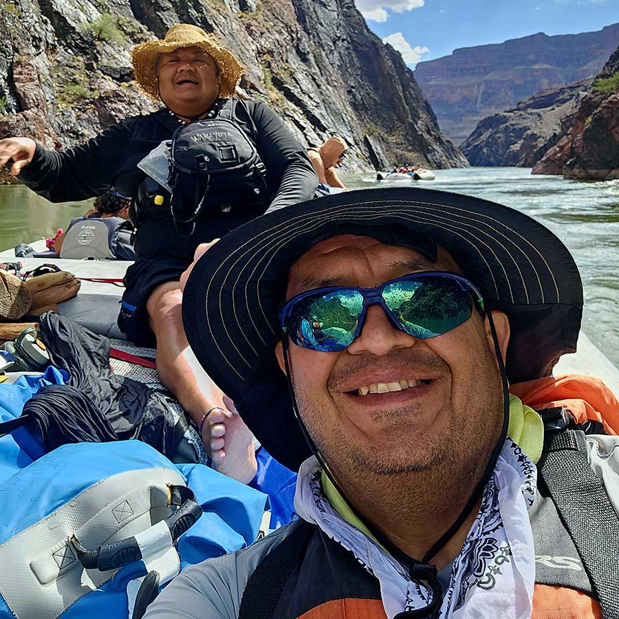 Two people wearing hats and sunglasses smile while rafting through a canyon river, surrounded by gear and rugged rock walls under a sunny sky.