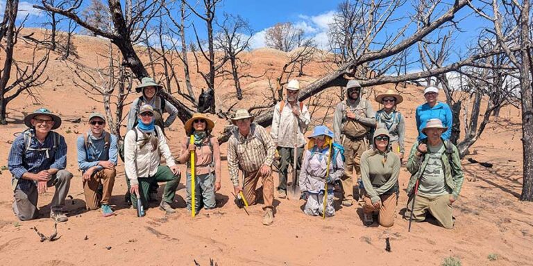 Group of people wearing hats and outdoor clothing pose in a dry, sandy landscape with sparse, leafless trees under a blue sky.