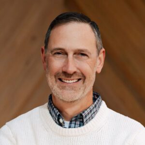 Chris Glick, a middle-aged man with short dark hair and a gray beard, is smiling while wearing a white sweater over a plaid shirt, standing in front of a wooden background.