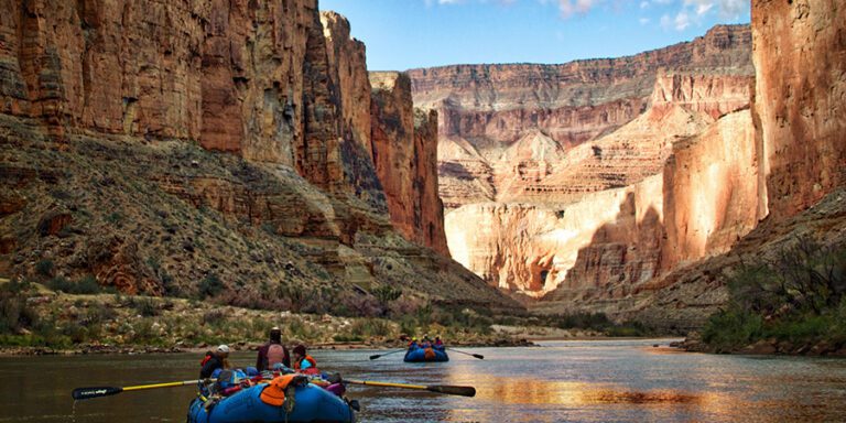 Several people in blue rafts paddle down the Colorado River in the Grand Canyon flanked by steep, sunlit canyon walls under a partly cloudy sky.