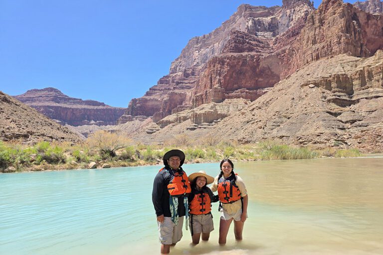 Three people wearing orange life vests and hats stand in shallow water on a Colorado River trip, with rocky canyon walls and blue sky in the background.