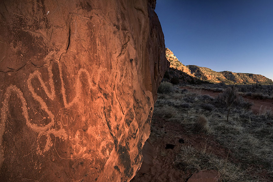 Petroglyphs carved into a sunlit red rock wall in a rugged, arid landscape and hills stretching beneath a clear sky.