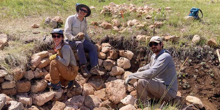 Three people wearing hats and work clothes build a stone wall outdoors on a grassy field, with rocks and tools scattered around.