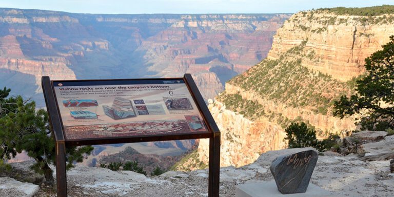Interpretive sign about Vishnu rocks stands at the edge of the Grand Canyon, with layered canyon walls and a rock sample visible in the background.