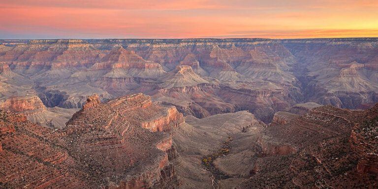 Grand Canyon landscape at sunrise, showing layered rock formations and deep valleys of the Grand Canyon from the South Rim, beneath a colorful sky.