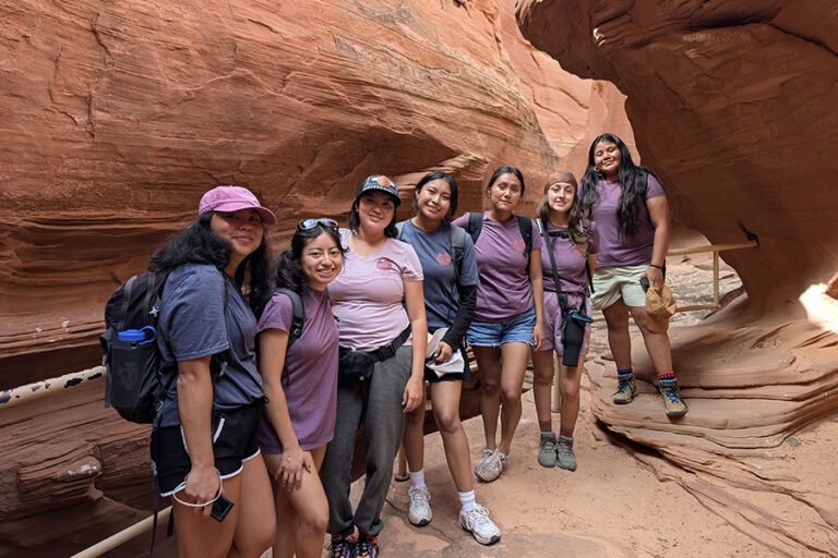 Seven young women pose together in a narrow, red rock canyon, wearing casual hiking attire and carrying backpacks as part of the 2025 LeaderShift Scholars Program.