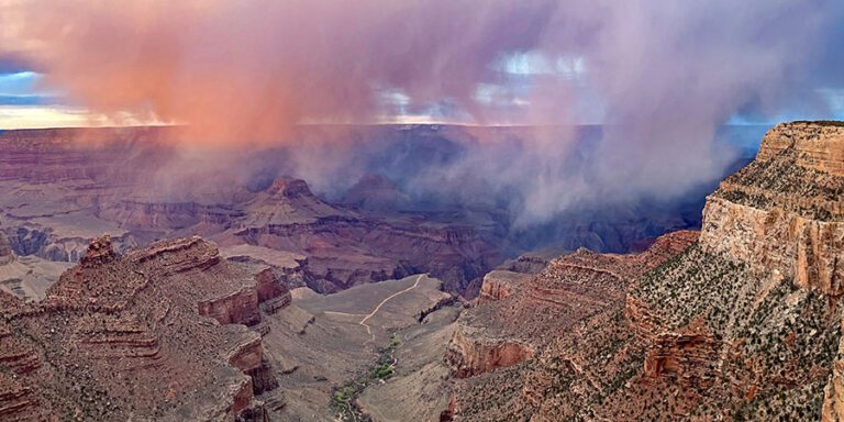 Colorful clouds and mist hover over the layered rock formations of the Grand Canyon at sunset, casting vibrant hues across the landscape.