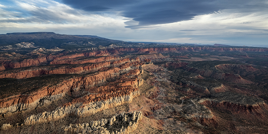 Aerial view of a vast, rugged desert landscape with layered red rock formations under a partly cloudy sky.