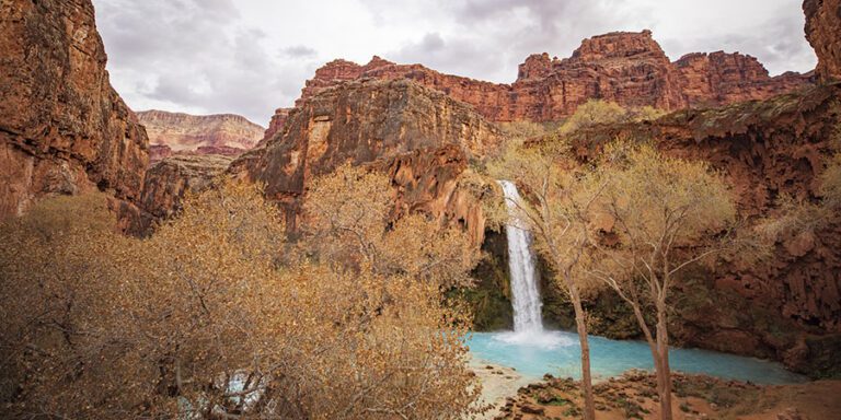 A waterfall flows into a turquoise pool surrounded by red rock cliffs and autumn trees under a cloudy sky.