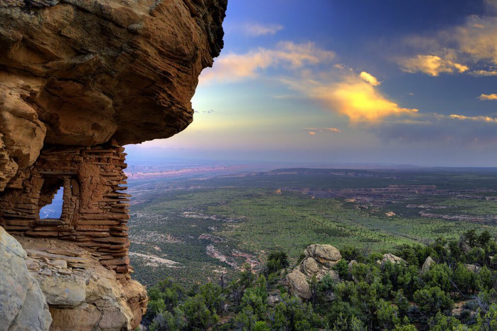 Ancient stone structure built into a cliff overlooks a vast green valley and distant mesas under a colorful evening sky.
