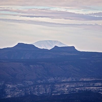 Distant mountain peak visible through a hazy sky, behind two flat-topped mesas and layered rock formations in the foreground; a landscape.