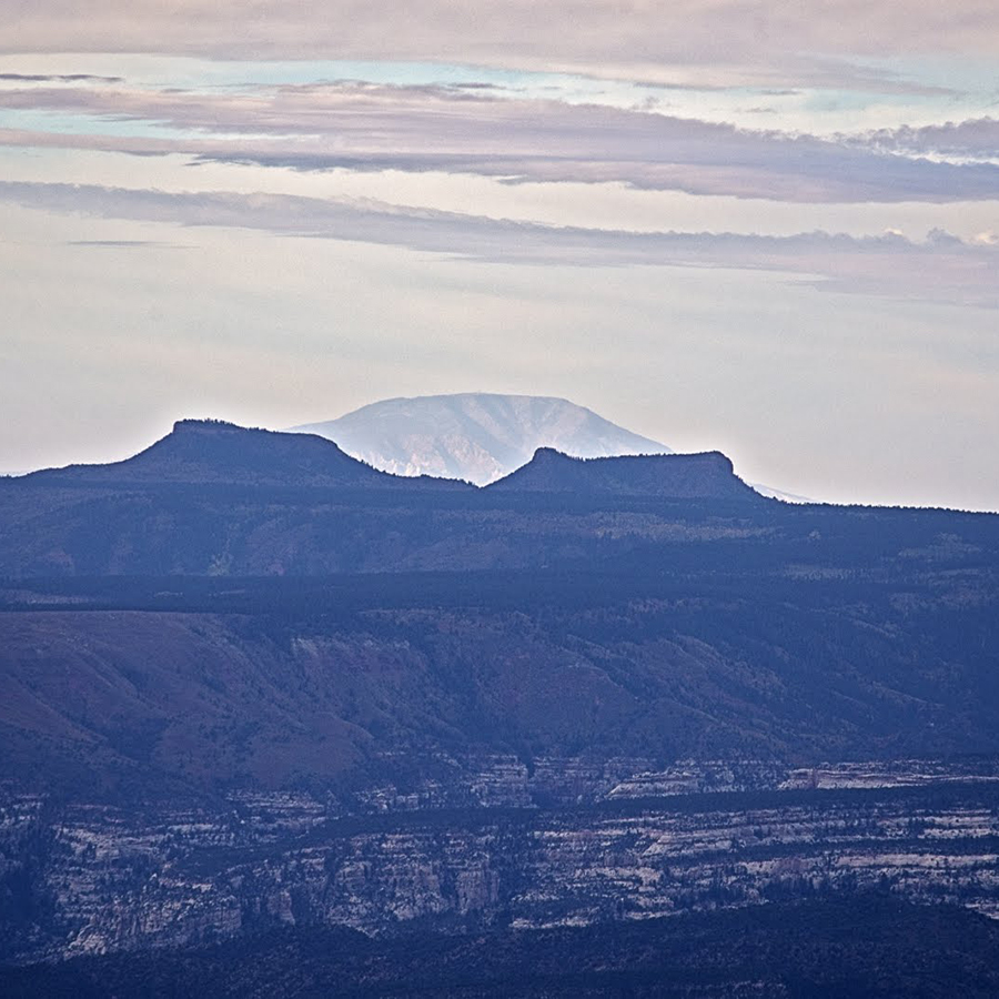Distant mountain peak visible through a hazy sky, behind two flat-topped mesas and layered rock formations in the foreground; a landscape.