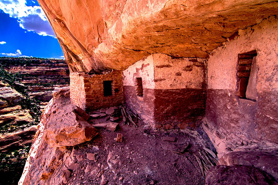 Ancient stone cliff dwelling built into a canyon wall, showing partial ruins with small windows and doorways, under an overhanging rock.