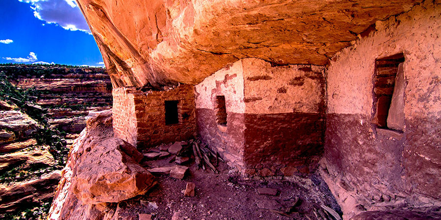 Ancient cliff dwelling built into a rocky canyon wall, with stone walls, small windows, and rubble on the ground under a bright sky—its preservation often highlighted in press releases celebrating historical discoveries.