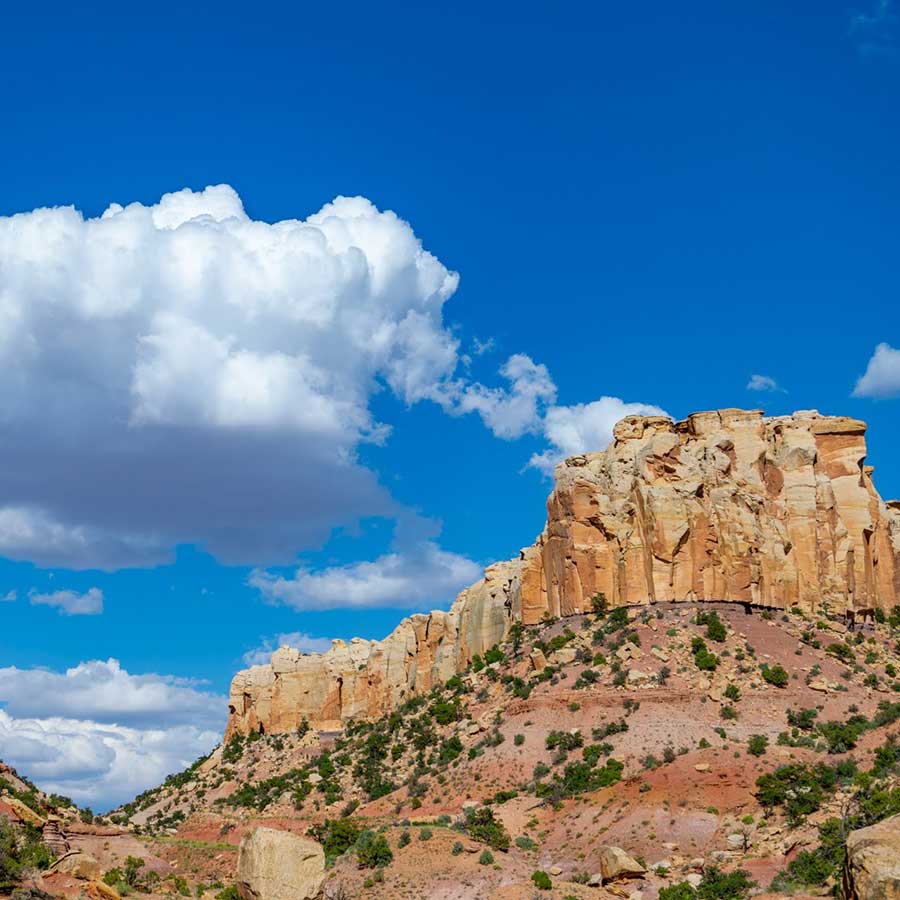 A rocky sandstone cliff with sparse vegetation under a bright blue sky with large, white clouds.