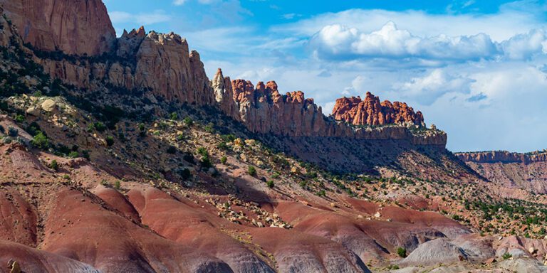 Rocky red hills and cliffs under a blue sky with scattered clouds, featuring sparse vegetation and dramatic geological formations in Grand Staircase-Escalante National Monument.