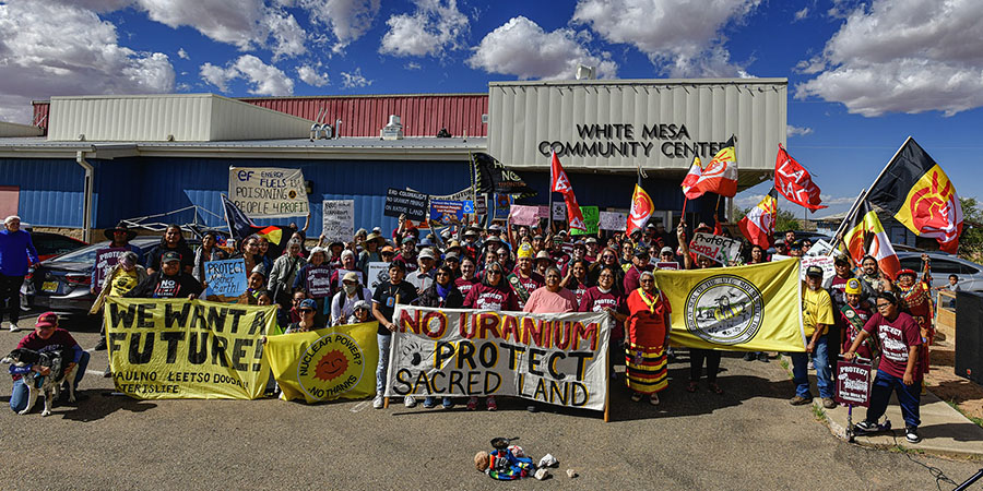 A group of people hold banners and signs protesting uranium mining by Energy Fuels in front of the White Mesa Community Center under a partly cloudy sky.