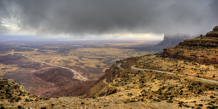 A winding road ascends a rocky, desert cliff under a cloudy sky, with a vast arid plain stretching into the distance.