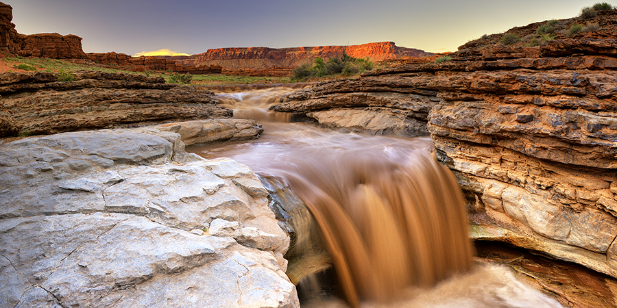 A muddy river flows over a rocky ledge, creating a small waterfall amid layered red rock formations under a clear sky at sunset.