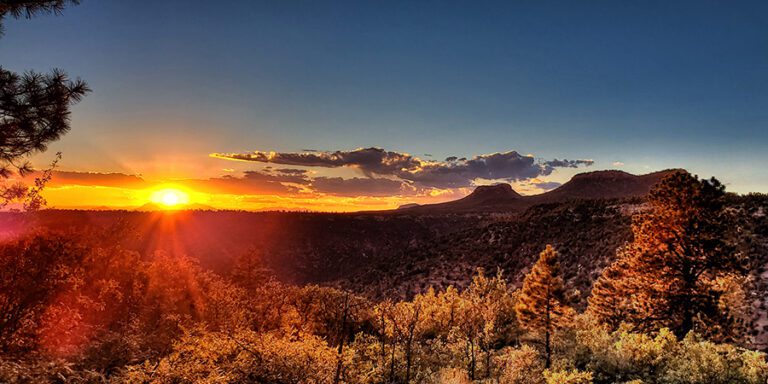 Sunset over a mountainous landscape with trees in the foreground, the sun near the horizon, and clouds lit by orange and yellow light.