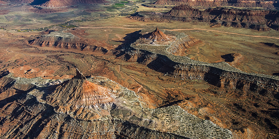 Aerial view of a desert landscape with red rock formations, mesas, and sparse vegetation, featuring rugged terrain and distant green patches.